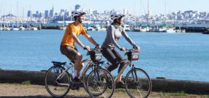 Two people riding tandem bike with San Francisco skyline in background